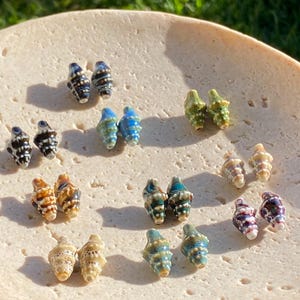 May include: A collection of small, conch-shaped ceramic beads arranged in pairs. The beads feature various glossy glaze colours including dark blue, light blue, green, brown, tan, and purple, each with textured, ridged surfaces and white detailing.