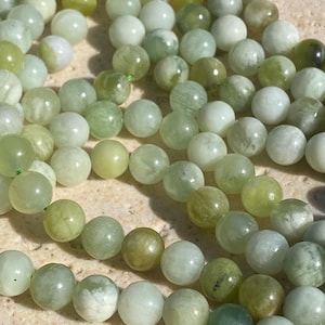 May include: A collection of round, smooth beads in various shades of light green, pale yellow, and translucent white. The beads are strung together on a thin green cord, displaying natural variations in colour, opacity, and internal mineral patterns.