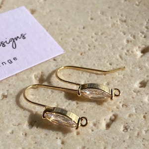 May include: A pair of gold drop earrings, each featuring a clear, oval-cut stone. The earrings are presented on a textured, pale surface. A white card with the word "signs" is partially visible. The earrings are likely for pierced ears.