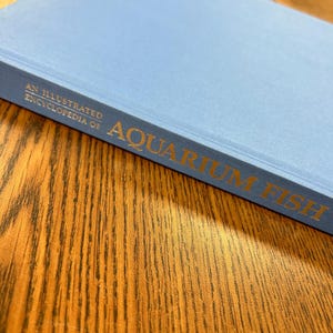 Può includere: Un libro rilegato in blu intitolato "An Illustrated Encyclopedia of Aquarium Fish".