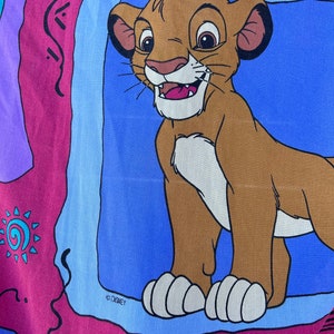 Puede incluir: Una tela colorida con una imagen de dibujos animados de Simba, el cachorro de león de El Rey León de Disney. La imagen está rodeada por un borde azul y el texto "© DISNEY" es visible en la esquina inferior izquierda.