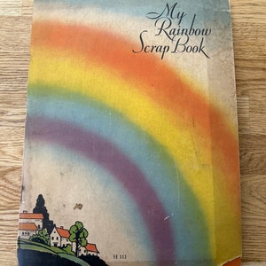 Może przedstawiać: Vintage album na zdjęcia zatytułowany "My Rainbow Scrap Book". Okładka przedstawia żywy łuk tęczy u góry, z malowniczą sceną wioski na dole. Paleta kolorów obejmuje odcienie niebieskiego, pomarańczowego, żółtego i fioletowego.