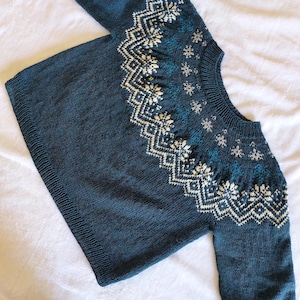 Peut inclure: Un pull tricoté bleu canard foncé avec un col rond et des manches longues. Le pull présente un motif géométrique crème et bleu canard sur la poitrine et les épaules, avec des motifs de flocons de neige. Le tricot est présenté sur une surface blanche.