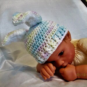 May include: A pastel-coloured crochet baby hat with bunny ears, featuring blue, green, and pink hues. The hat is on a baby doll. The doll wears a yellow shirt and rests on a white surface.
