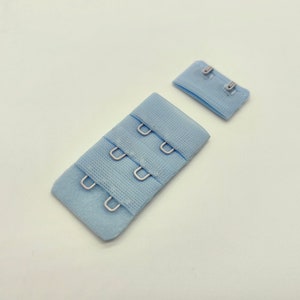 Könnte beinhalten: Zwei hellblaue BH-Verlängerungen mit Metallhaken. Die Verlängerung links hat drei Reihen Haken, während die Verlängerung rechts zwei Reihen Haken hat.