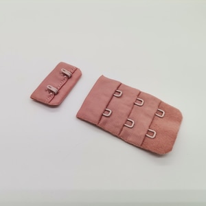 Può includere: Un set di due chiusure a gancio e occhiello per reggiseno rosa. La chiusura a sinistra ha tre ganci, e la chiusura a destra ha tre file di ganci.