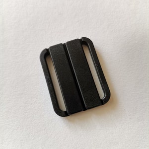 May include: Black plastic slider buckle with two parallel slots. The buckle has the word "KSM" printed on it.