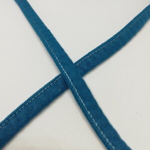 May include: Two pieces of teal blue fabric are sewn together to form a cross shape. The fabric has a white stitching detail along the edges.