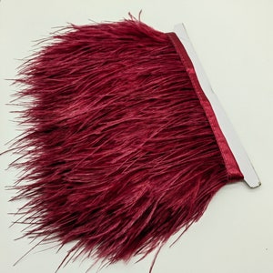 Pode incluir: Uma guarnição de penas de avestruz de cor bordô com um suporte de cetim. A guarnição tem aproximadamente 15 cm de largura e é perfeita para adicionar um toque de elegância a qualquer projeto.