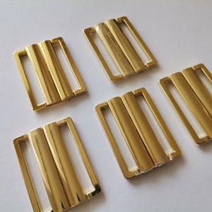 Op de afbeelding: Vijf goudkleurige metalen gespen, elk met twee parallelle staven, ontworpen om banden of stof te bevestigen. Deze gespen worden vaak gebruikt bij het maken van kleding en accessoires.