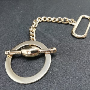 Puede incluir: Una cadena de metal dorado con un anillo redondo y un cierre de palanca. El anillo tiene un dise&ntilde;o decorativo con un peque&ntilde;o pomo redondo en un lado.