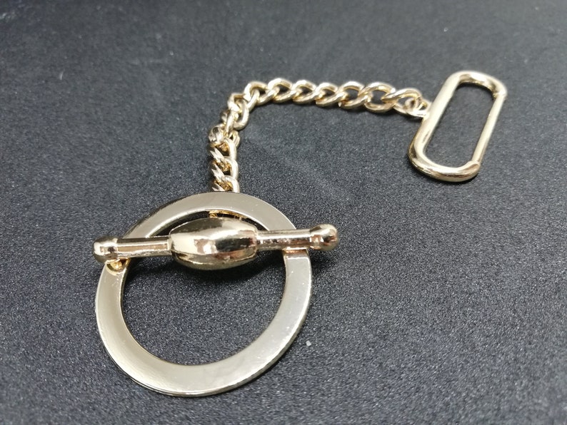 Puede incluir: Una cadena de metal dorado con un anillo redondo y un cierre de palanca. El anillo tiene un dise&ntilde;o decorativo con un peque&ntilde;o pomo redondo en un lado.