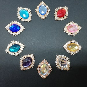 Könnte beinhalten: Sortiment aus zehn ovalen, juwelenbesetzten Verzierungen mit goldenen Fassungen. Jede Verzierung hat einen andersfarbigen Edelstein, darunter blau, rot, rosa, lila und klar.