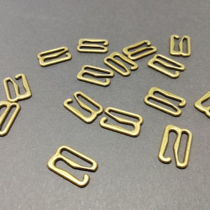 Op de afbeelding: Een groep van 15 goudkleurige metalen bh-bandverstellers, ook wel bh-haken genoemd, liggen verspreid op een grijs oppervlak.