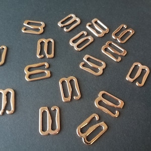 Könnte beinhalten: Eine Gruppe von 15 goldfarbenen Metall-BH-Gurt-Verstellern. Die Versteller sind S-förmig und haben an jeder Seite zwei Schlaufen.