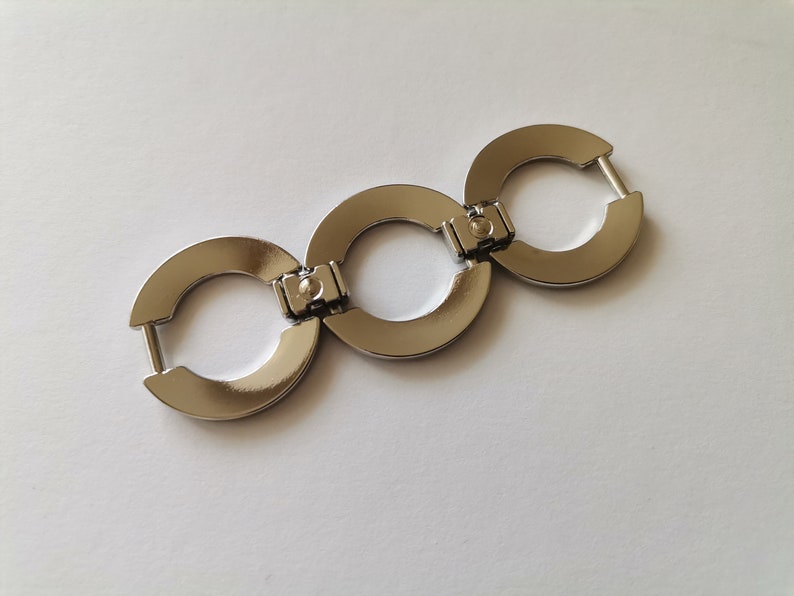 May include: Three silver metal rings connected together, forming a chain. Each ring has a smooth, rounded surface and a small, circular opening in the center.