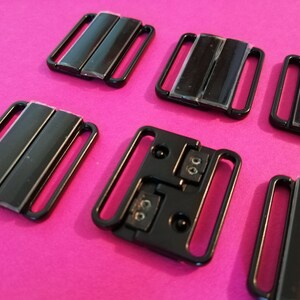 Puede incluir: Seis cierres de sujetador de pl&aacute;stico negro, algunos con dos filas de metal, otros con una sola fila. Los cierres est&aacute;n sobre una superficie rosa.