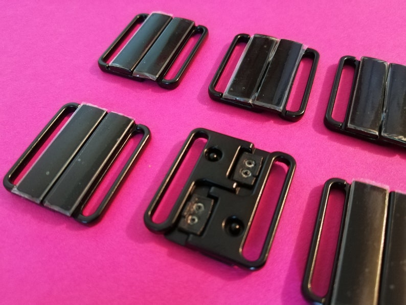 Puede incluir: Seis cierres de sujetador de pl&aacute;stico negro, algunos con dos filas de metal, otros con una sola fila. Los cierres est&aacute;n sobre una superficie rosa.