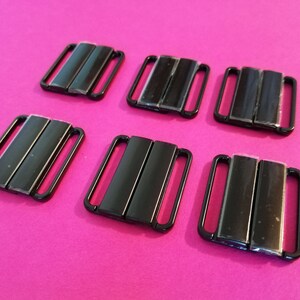 Puede incluir: Seis cierres de sujetador de metal negro, tambi&eacute;n conocidos como ganchos de sujetador, est&aacute;n dispuestos en dos filas de tres sobre una superficie rosa.