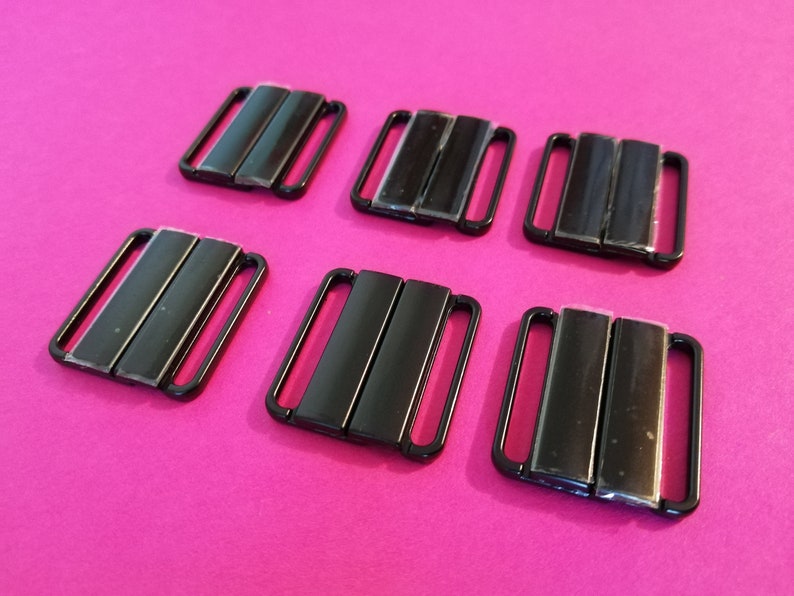 Puede incluir: Seis cierres de sujetador de metal negro, tambi&eacute;n conocidos como ganchos de sujetador, est&aacute;n dispuestos en dos filas de tres sobre una superficie rosa.