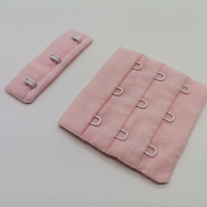 May include: Two pink fabric bra hook and eye closures. The larger closure has three rows of hooks and eyes. The smaller closure has three hooks and eyes.