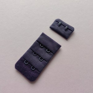 Pode incluir: Dois extensores de sutiã de tecido preto com ganchos de metal. O extensor da esquerda tem quatro ganchos e o extensor da direita tem dois ganchos.