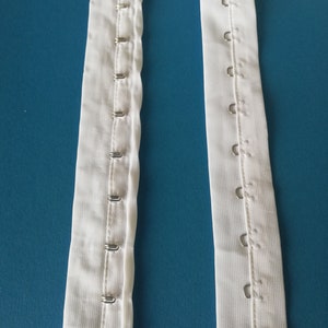 Puede incluir: Dos tiras de tela blanca con filas de ganchos met&aacute;licos cosidos a lo largo de los bordes. Los ganchos est&aacute;n dise&ntilde;ados para sujetarse a una prenda u otra tela.