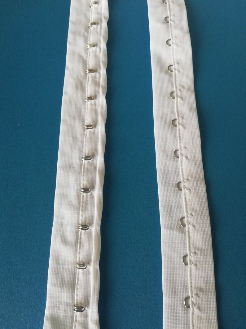 Puede incluir: Dos tiras de tela blanca con filas de ganchos met&aacute;licos cosidos a lo largo de los bordes. Los ganchos est&aacute;n dise&ntilde;ados para sujetarse a una prenda u otra tela.