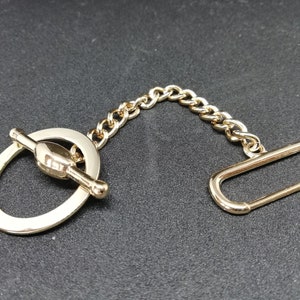 Puede incluir: Una cadena de metal dorado con un cierre de palanca y un anillo en un extremo y un bucle rectangular en el otro extremo.