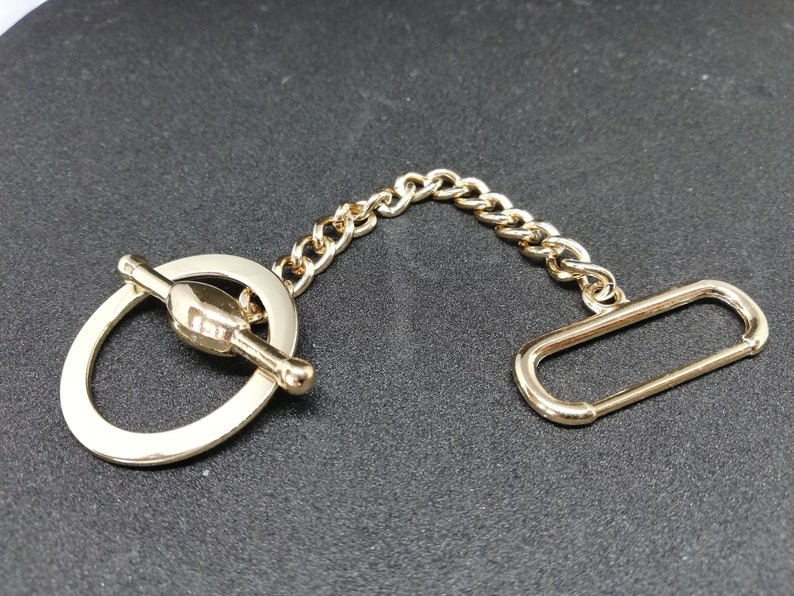 Puede incluir: Una cadena de metal dorado con un cierre de palanca y un anillo en un extremo y un bucle rectangular en el otro extremo.