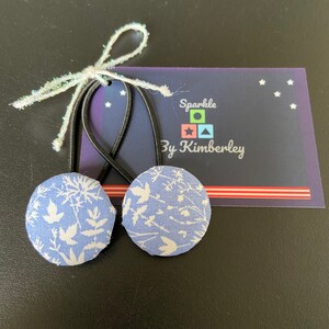May include: Two blue fabric hair scrunchies with a white floral pattern. The scrunchies are tied together with a white ribbon. The background is black with a white card that says "Sparkle By Kimberley" with a colourful geometric design.