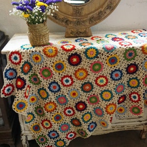 May include: A cream-colored, crocheted table runner with a repeating pattern of colorful floral motifs. The flowers are in shades of red, yellow, blue, green, and orange. The runner is draped over a white, distressed cabinet.