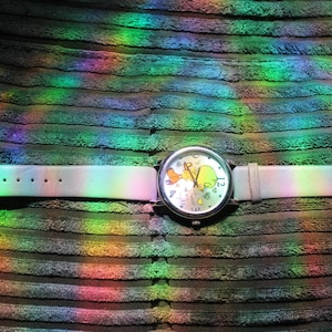 May include: A wristwatch with a light-colored band and a silver-toned case. The watch face features a cartoon character design. The watch band is approximately 9 inches long. The watch is set against a textured, dark background with rainbow light reflections.