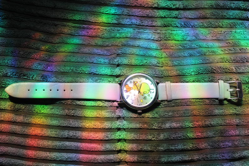 May include: A wristwatch with a light-colored band and a silver-toned case. The watch face features a cartoon character design. The watch band is approximately 9 inches long. The watch is set against a textured, dark background with rainbow light reflections.