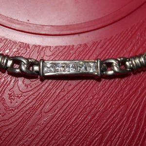 May include: A silver-toned metal chain necklace featuring a central rectangular bar link embedded with four clear, square-cut stones. The chain consists of alternating smooth oval links and textured, ribbed cylindrical segments throughout the design.