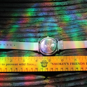 May include: A wristwatch with a pearlescent band and a silver-colored watch face. The watch is laid out next to a yellow ruler with black markings and text that reads "A rule to prevent wild fires..." and "Smokey's Friends Don't Play With Fire."