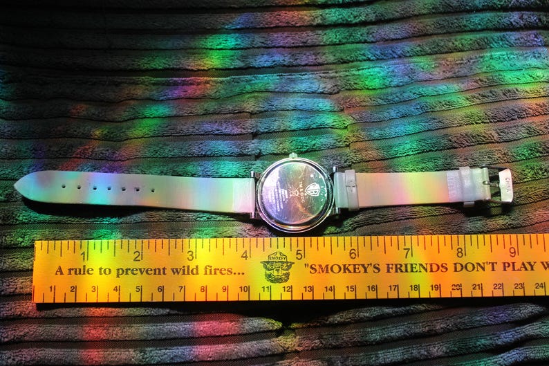 May include: A wristwatch with a pearlescent band and a silver-colored watch face. The watch is laid out next to a yellow ruler with black markings and text that reads "A rule to prevent wild fires..." and "Smokey's Friends Don't Play With Fire."