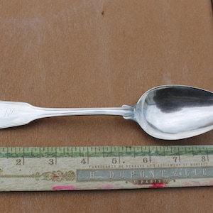 May include: A silver spoon with a rounded bowl and a decorative handle. The handle has a heart-shaped design and an engraved monogram. A vintage ruler with inch measurements is below the spoon.