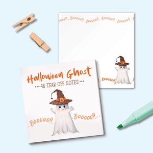 May include: A stack of square paper notes featuring a white ghost wearing a brown witch hat with an orange band. The text reads Halloween Ghost 48 Tear Off Notes and Booooo. Two wooden clothespins and a light green marker are positioned nearby.