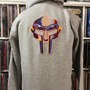Op de afbeelding: Een grijze hoodie met een kleurrijk geborduurd maskerontwerp op de rug. Het masker heeft tinten paars, oranje en blauw. De hoodie heeft een capuchon met trekkoord en lange mouwen.