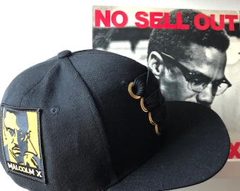 May include: Black baseball cap with a yellow and black patch featuring a portrait of Malcolm X and the text "Malcolm X". The cap has a black lace-up detail with gold eyelets.