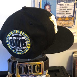 Puede incluir: Gorra de béisbol negra con un logotipo blanco y un parche que dice "ODB BASTARD 1968-2004". La gorra se muestra con un cinturón negro con una hebilla dorada que dice "THC". También es visible una cadena dorada.