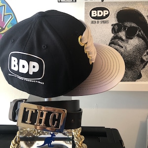 May include: Black baseball cap with a white "BDP" logo and gold accents. The cap has a beige brim and is sitting on top of a brown leather belt with a silver buckle that says "THC". A gold chain is wrapped around the belt.