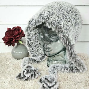 May include: A gray and white faux fur hat with a long, fluffy brim and matching ties. The hat is displayed on a mannequin head.