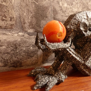 Puede incluir: Una escultura oscura y texturizada de una criatura sosteniendo una esfera naranja con detalles rojos y amarillos. La escultura tiene un acabado rugoso y moteado y está colocada contra una pared de piedra y una superficie de madera.