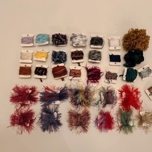 May include: Assortment of colorful yarn spools and fluffy yarn balls in various colors, including red, blue, green, yellow, brown, and white. The yarn is arranged in rows on a white surface.