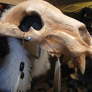 Puede incluir: Una réplica de cráneo animal beige sobre una base de tela azul oscuro, adornada con un pendiente de cadena plateado. El pendiente incluye un pequeño perno circular, dos cuentas ovaladas oscuras y una pluma larga de color claro.