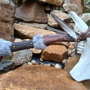 Peut inclure: Hache décorative artisanale avec une lame en os et un manche en bois. Le manche est enveloppé de cuir marron, orné de fourrure grise et de plumes. L'hache est présentée sur un fond de roches naturelles.