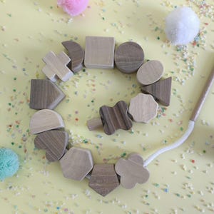 Pode incluir: Colar de contas de madeira com várias formas geométricas em tons de madeira natural. O colar está sobre uma superfície amarela com confetes coloridos e pompons fofos em rosa, branco e azul-petróleo.