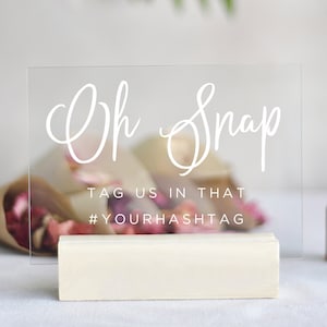 May include: A clear acrylic sign with white text that reads "Oh Snap" and "Tag us in that #YOURHASHTAG". The sign is on a small wooden stand.